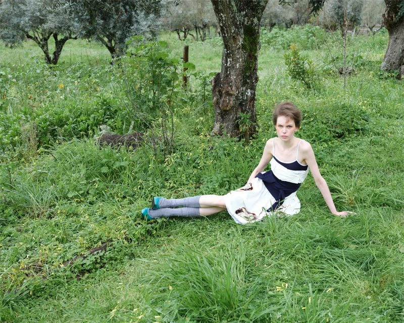 A slender young woman with short hair sits on a grassy field surrounded by trees, wearing a summer dress and socks with teal flats.