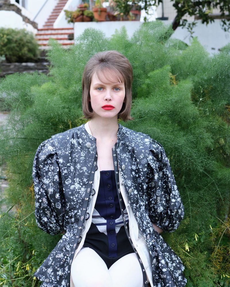 A fair-skinned woman with a retro hairstyle and red lipstick poses in a patterned jacket in front of a lush green bush outdoors.