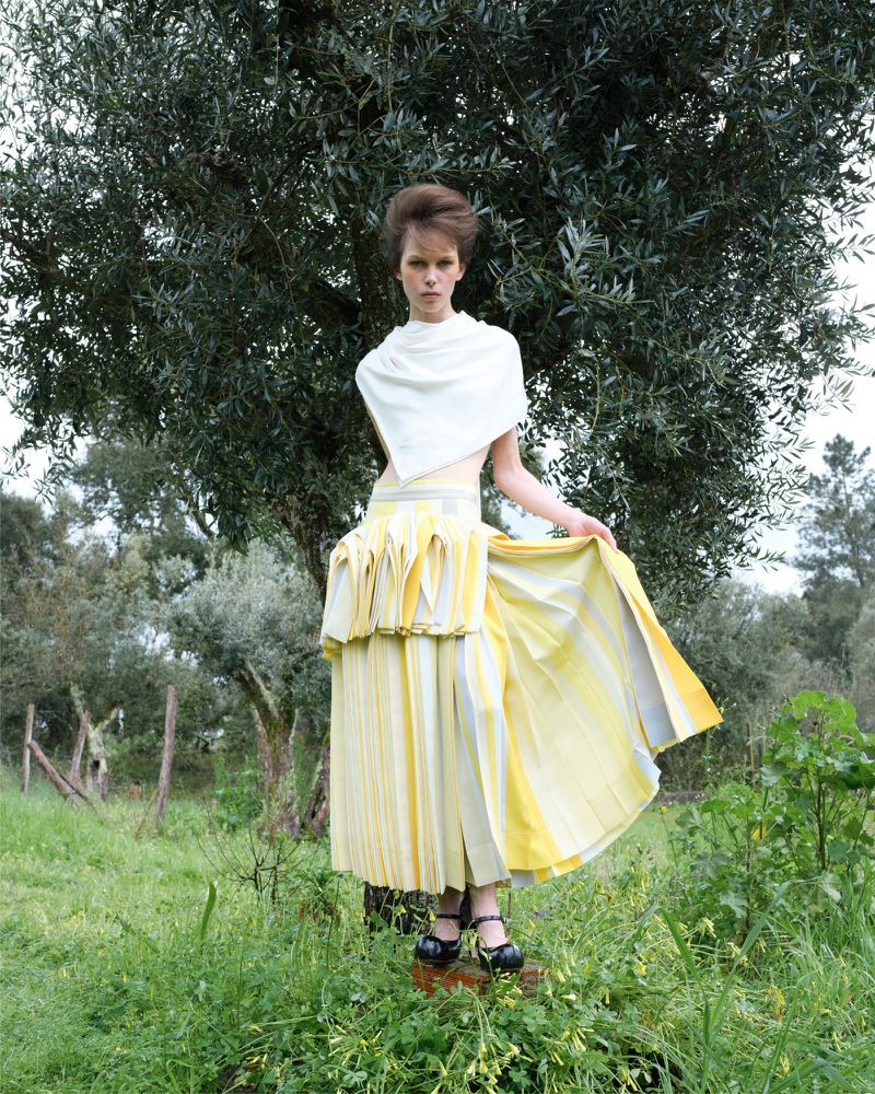 A slender woman models a layered yellow skirt and white shawl in a grassy outdoor setting, posing in front of a large, leafy green tree.