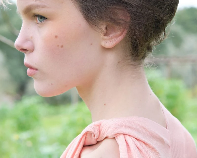 Profile shot of a fair-skinned young woman with light hair, gazing left in an outdoor setting, wearing a pink draped top.