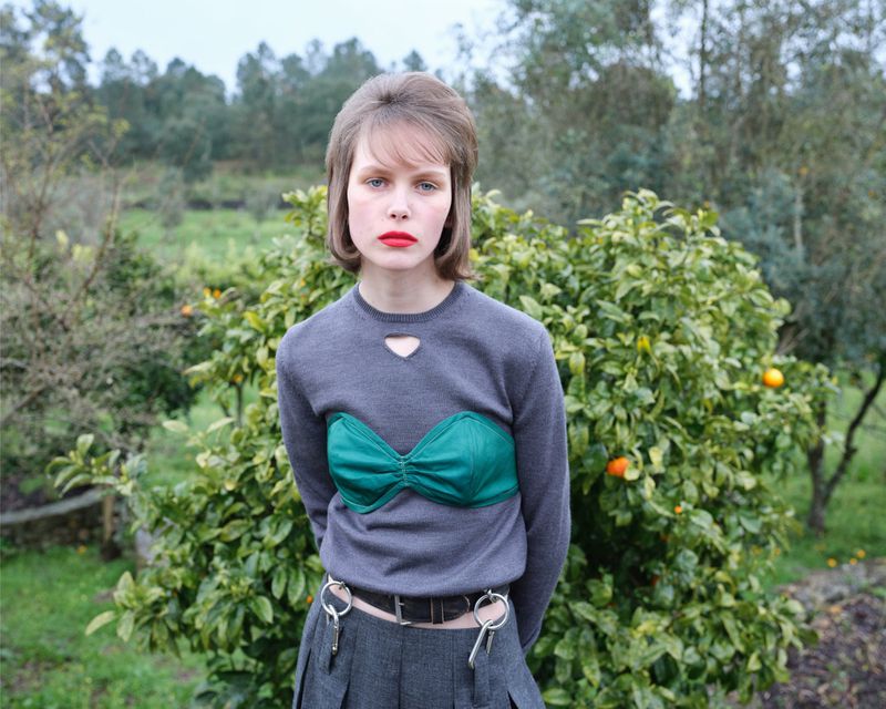 Portrait of a young woman with red lipstick wearing a gray sweater with a green bustier top, standing outside among orange trees.