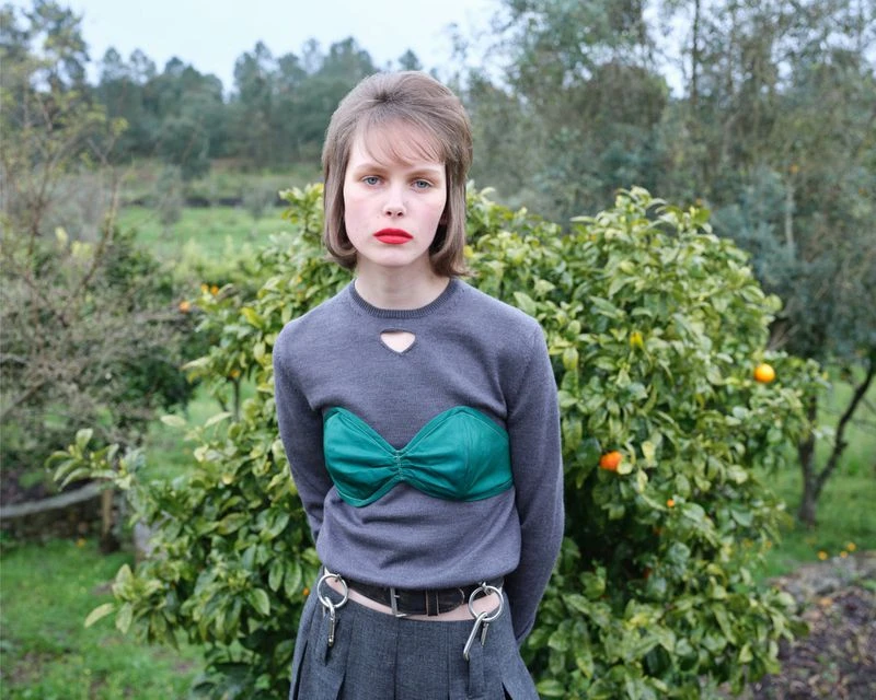 Portrait of a young woman with red lipstick wearing a gray sweater with a green bustier top, standing outside among orange trees.