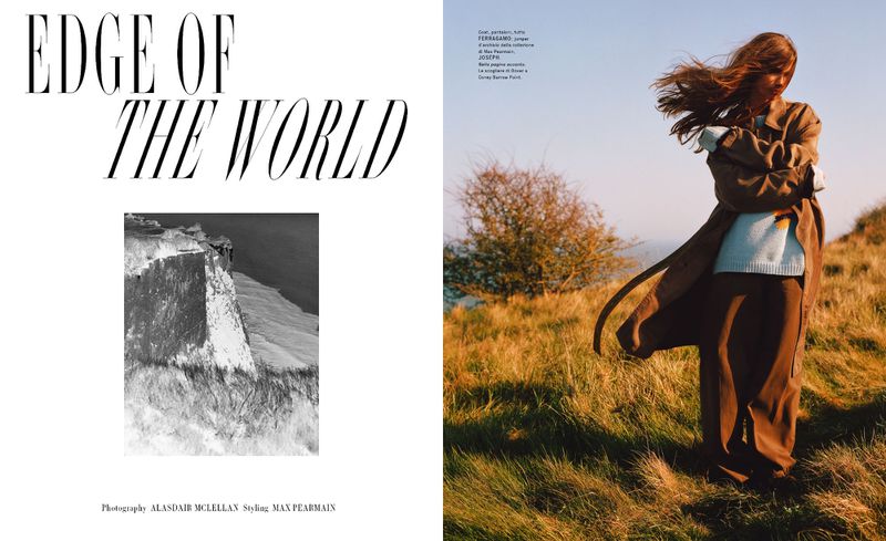 Fashion editorial photo of a woman in a long coat and dress on a grassy hilltop, hair blowing in the wind on a sunny day.