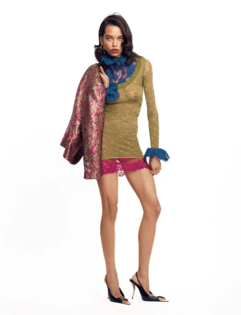 A fashionable woman posing in a studio. She wears a green patterned shirt, a pink lace skirt, and black slingback heels against white background.
