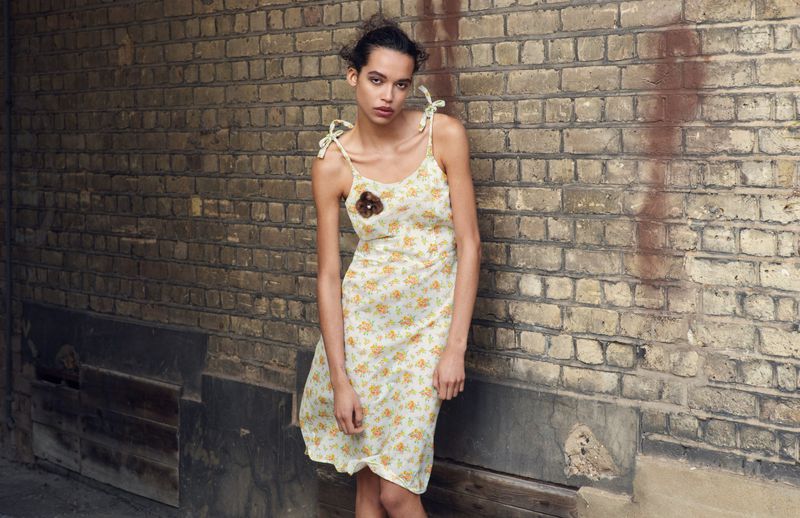 A young woman in a summery dress stands against a brick wall, captured in a medium shot showcasing the dress and neutral backdrop.