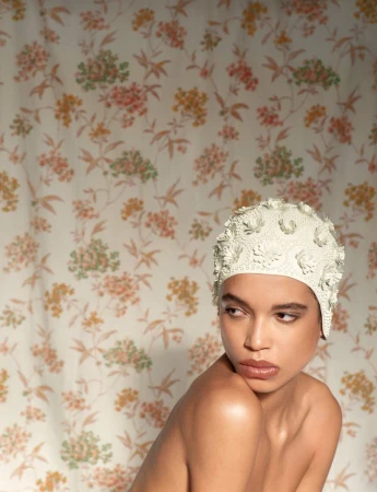 Portrait of adult female with bare shoulders and swimming cap, posing in front of a floral wallpaper background, indoor setting.