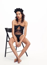 Model Lara wearing a black bodysuit and scalloped hat, seated on a black chair against a white background.
