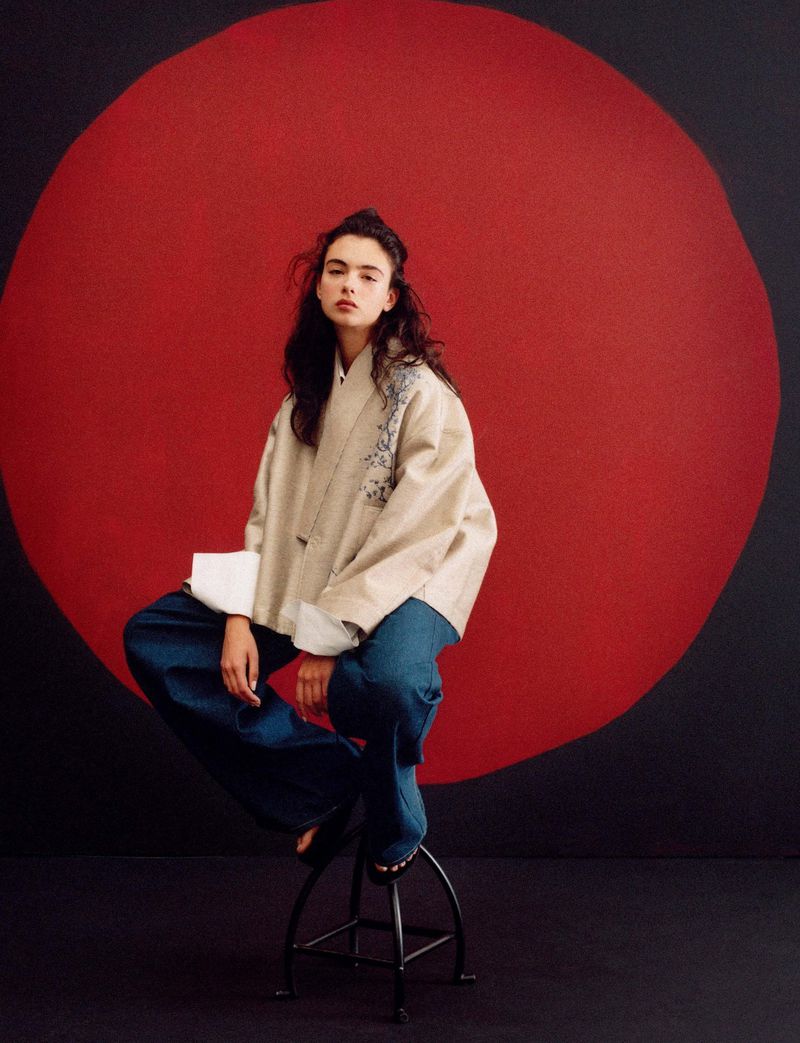 A stylish young woman in jeans and a neutral jacket sits on a stool against a graphic red and black backdrop in a studio setting.
