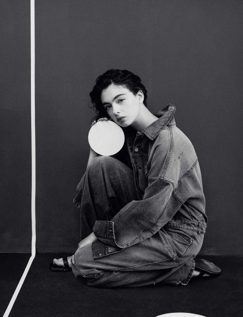 Black-and-white fashion portrait of a woman in a denim jumpsuit, seated with legs crossed, holding a white circular object against a dark backdrop.