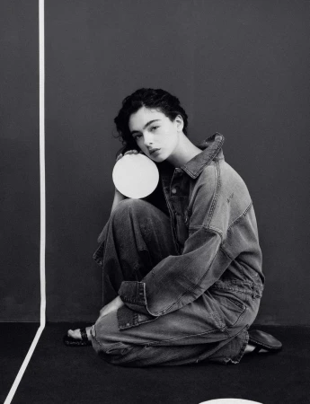 Black-and-white fashion portrait of a woman in a denim jumpsuit, seated with legs crossed, holding a white circular object against a dark backdrop.
