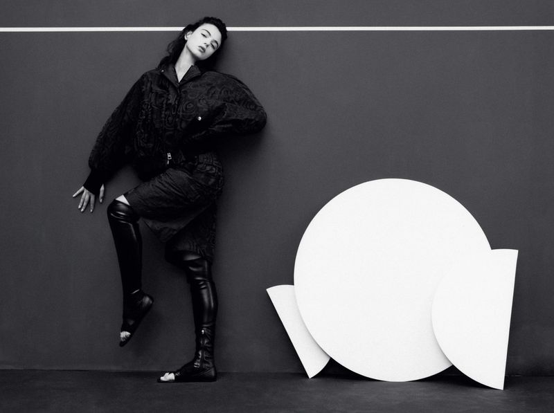 Fashionable woman in jacket and open-toe thigh-high boots poses against a gray wall with an abstract white circular geometric sculpture.
