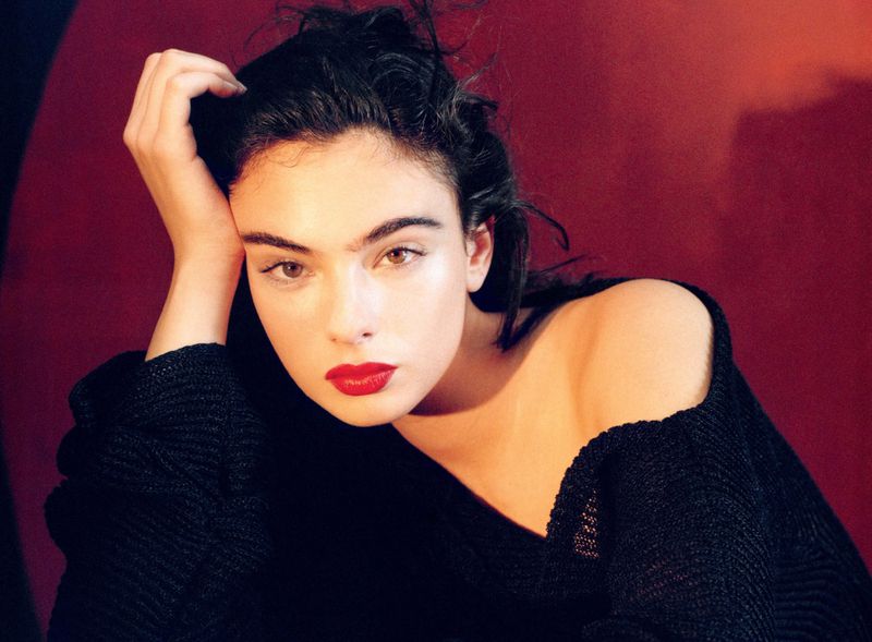 Close-up portrait of a woman with red lipstick and dark hair, set against a deep red background, creating a striking visual contrast.