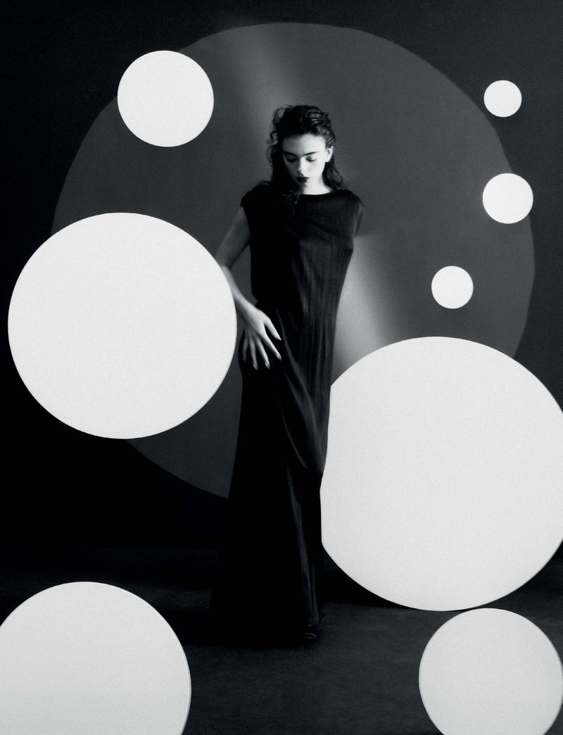 Elegant woman in a black dress poses in a studio surrounded by white circle props. Black and white studio fashion photography.
