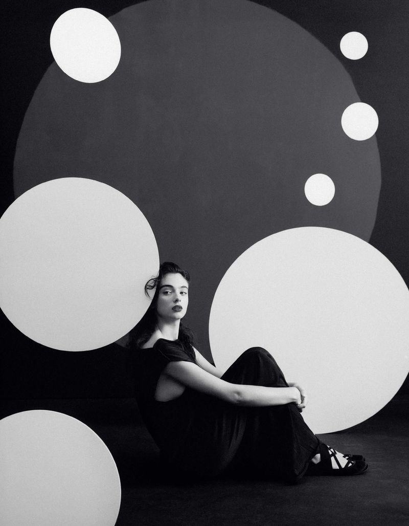 Monochrome portrait of a woman in a black dress sitting in front of a dark backdrop with white circular designs.