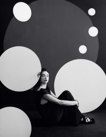 Monochrome portrait of a woman in a black dress sitting in front of a dark backdrop with white circular designs.