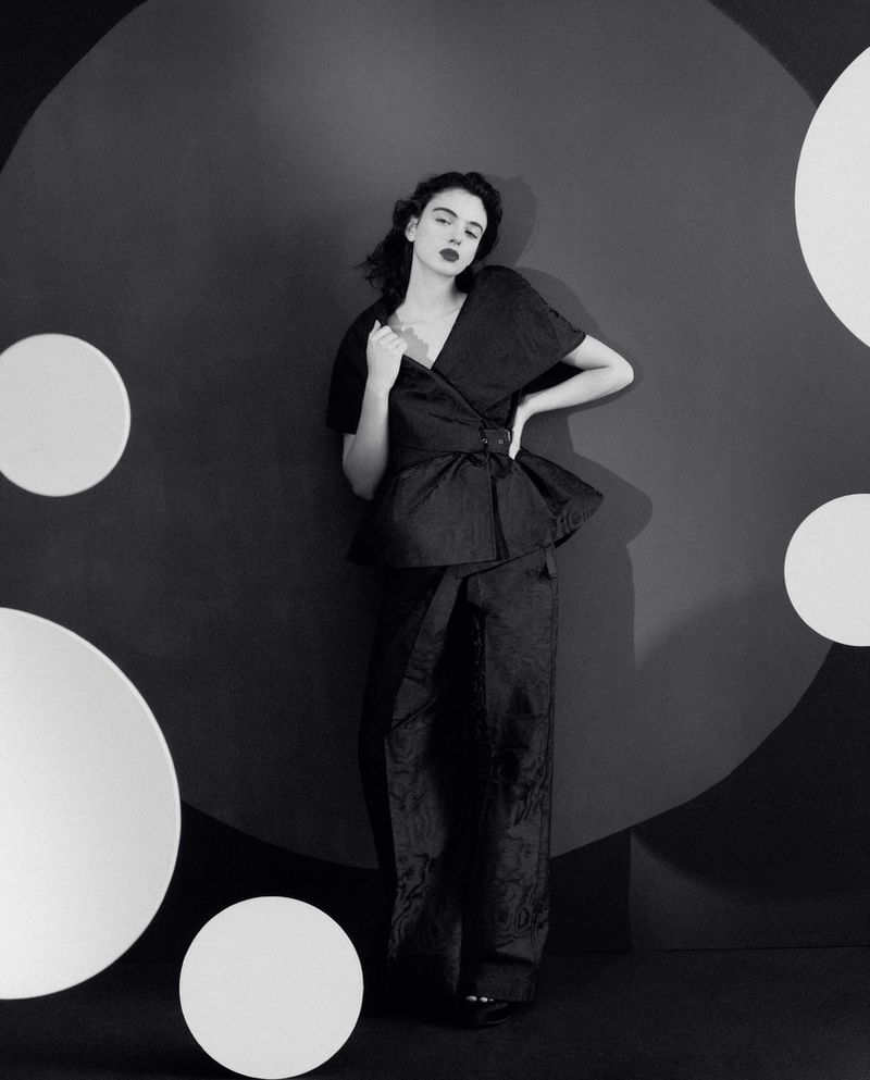 A woman in a dark, two-piece outfit poses in front of a gray wall with floating white circles in a studio fashion shot.