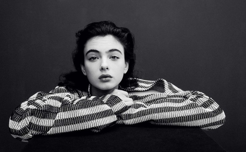 Black and white portrait of a woman with dark hair wearing a patterned top, resting her arms on a black surface.