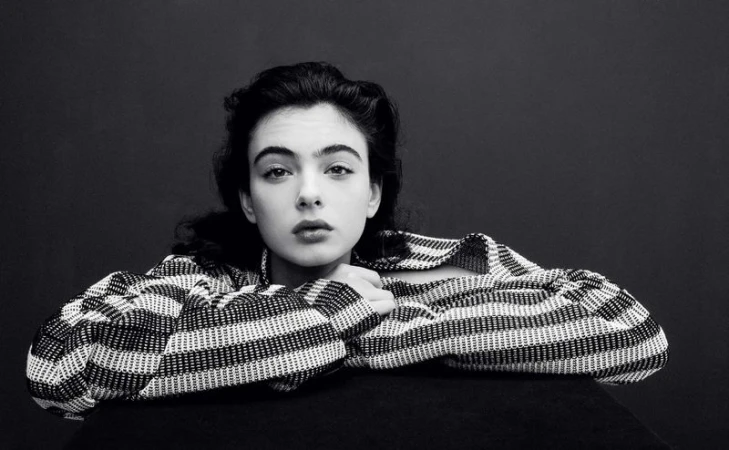 Black and white portrait of a woman with dark hair wearing a patterned top, resting her arms on a black surface.