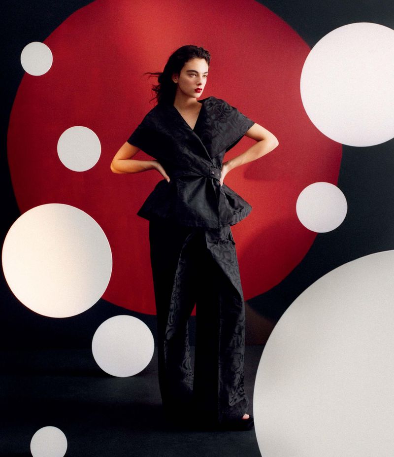A woman in a black brocade wrap top and wide-leg pants poses against a red and black backdrop with large white orb lights.