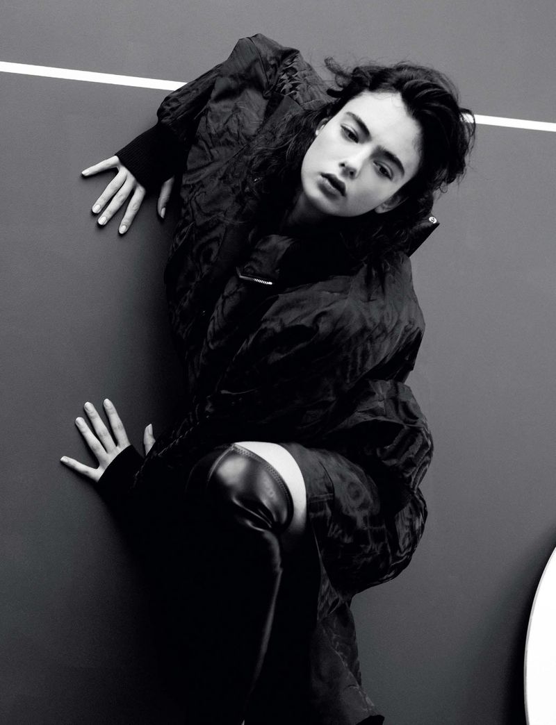 A woman in a quilted black coat and thigh-high boots poses in a studio with a black wall and a white line.