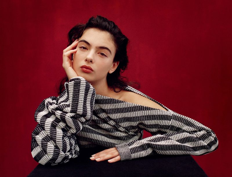 Portrait of a woman with dark hair in a black and white striped top against a red background. She has one hand on her face.