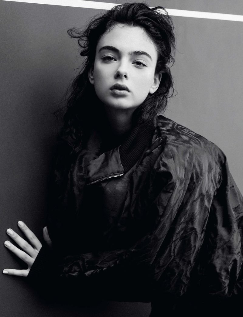 Monochrome portrait of a young adult woman with dark hair in a jacket against a dark gray background with a white stripe at the top.