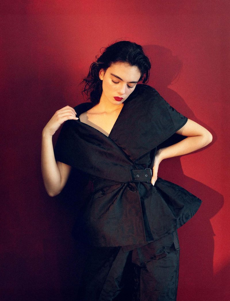Elegant woman in stylish black couture dress poses against a rich red backdrop in a fashion-forward portrait, showcasing texture and form.