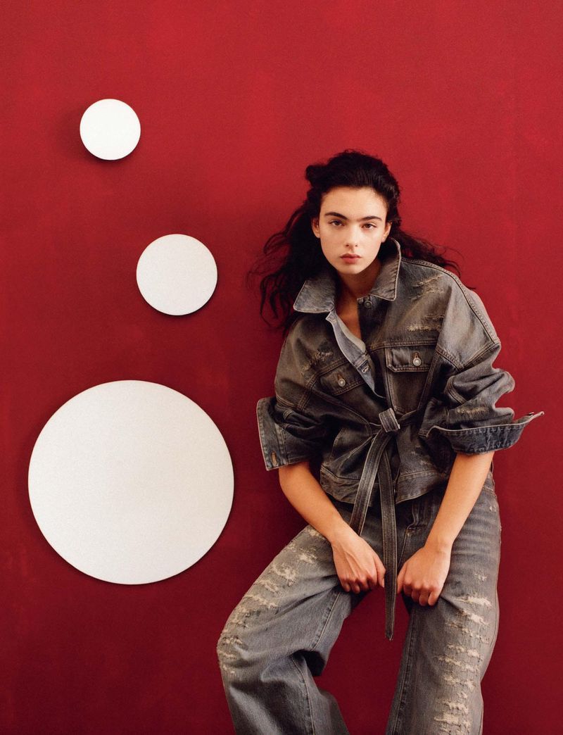 A woman poses indoors with dark hair wearing a denim jacket and ripped jeans, illuminated by indoor lighting against a red wall.