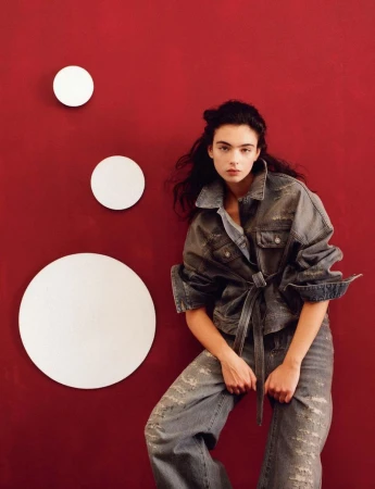 A woman poses indoors with dark hair wearing a denim jacket and ripped jeans, illuminated by indoor lighting against a red wall.