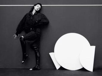 Model Deva Cassel in dark patterned jacket and knee-high boots posing against a dark wall with white circular geometric set props.