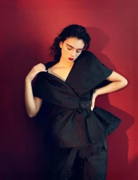 Model Deva Cassel wearing a structured black belted top against a dark red backdrop for Re-Edition Magazine