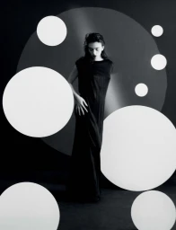 Deva Cassel in a dark sleeveless dress standing against a black background with overlaid white circular graphic elements
