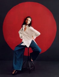 Model Deva Cassel seated on a stool wearing a white shirt and denim jeans against a red and black circular backdrop