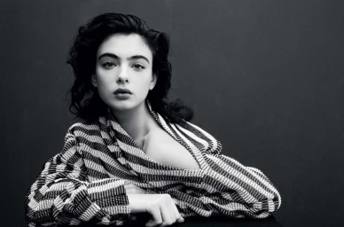 Monochrome portrait of model Deva Cassel wearing a patterned striped jacket against a dark background.
