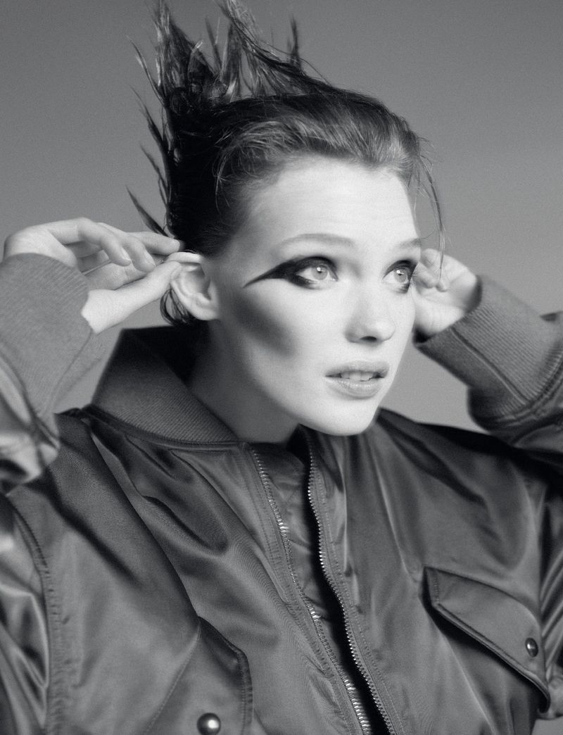 Black and white portrait of a young adult woman with striking eye makeup wearing a bomber jacket and styled hair, against a grey background.