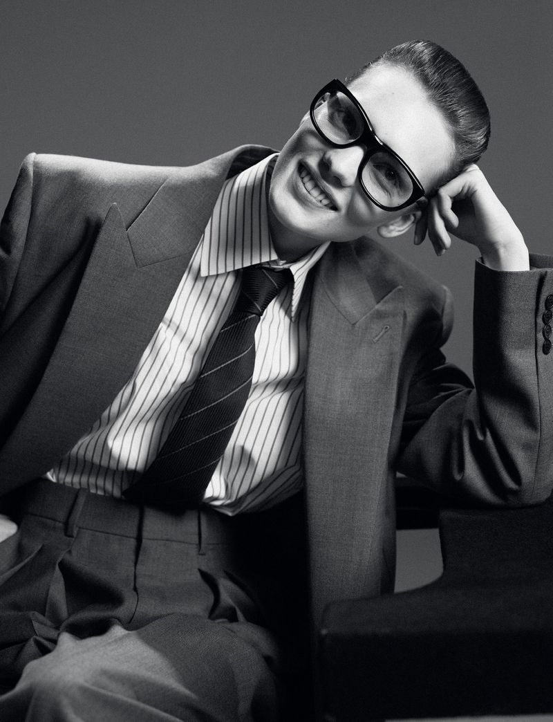 Black and white portrait of a smiling adult wearing a suit, pinstripe shirt, tie, and glasses, with hand raised to their head.