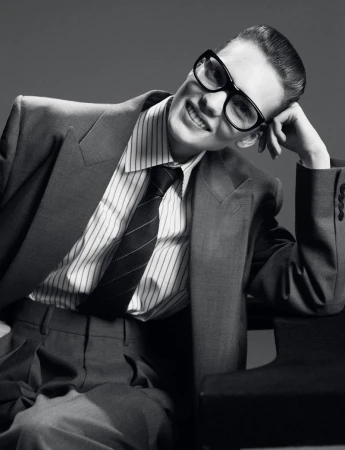 Black and white portrait of a smiling adult wearing a suit, pinstripe shirt, tie, and glasses, with hand raised to their head.