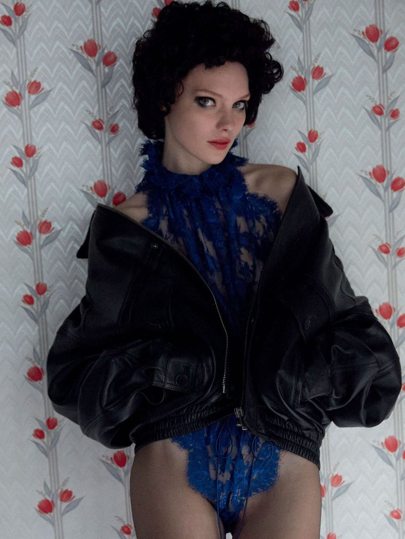 A woman with dark curly hair poses in blue lace lingerie and a black leather jacket against a patterned wallpaper backdrop indoors.