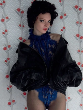 A woman with dark curly hair poses in blue lace lingerie and a black leather jacket against a patterned wallpaper backdrop indoors.