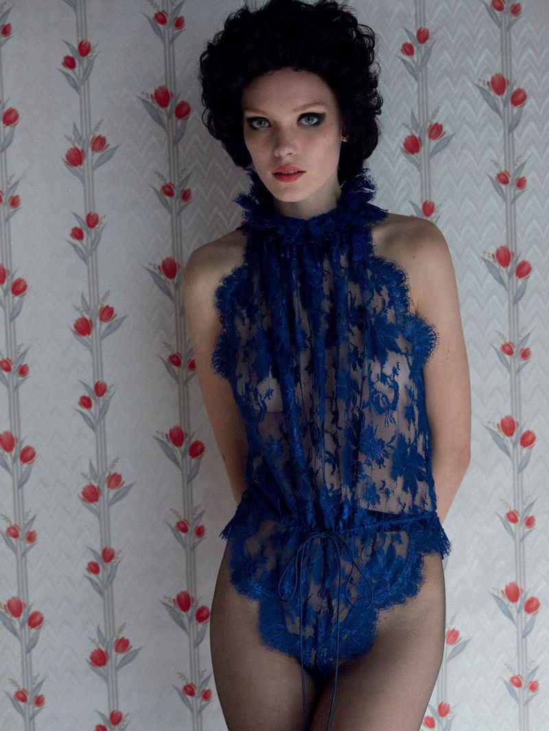 A young woman stands indoors against a floral wallpaper wearing a blue lace bodysuit, frilled high neck, and dark stockings.