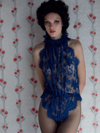 A young woman stands indoors against a floral wallpaper wearing a blue lace bodysuit, frilled high neck, and dark stockings.