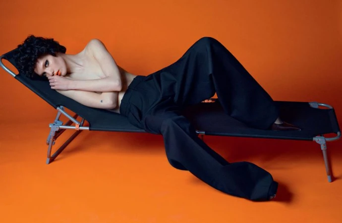 A woman with dark curly hair lounges on a black cot against an orange backdrop, wearing wide-leg black pants and minimal makeup.