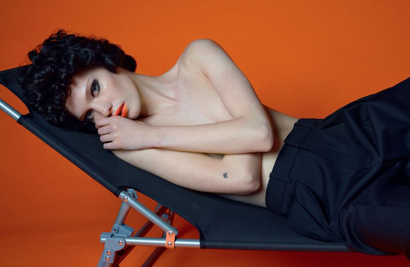 A woman with dark curly hair lies in a lounge chair with an orange backdrop, creating a striking contrast and a modern, relaxed aesthetic.