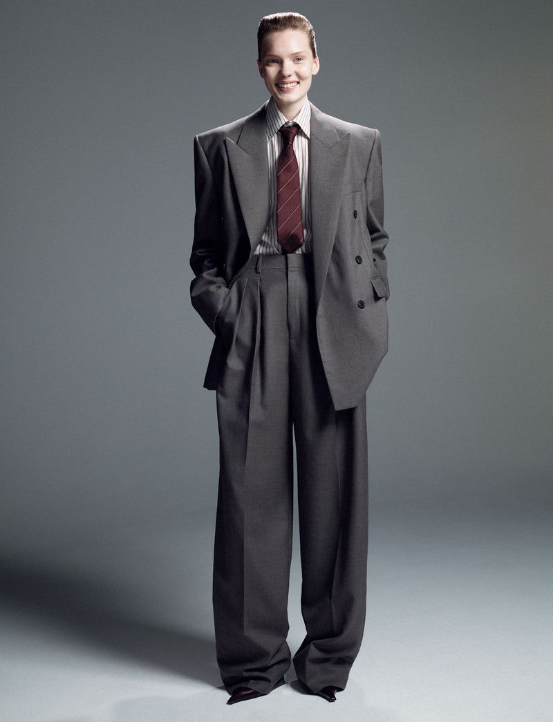 Smiling woman in an oversized grey suit with wide-leg pants, posing indoors against a neutral grey backdrop in a fashion editorial.