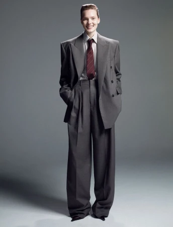 Smiling woman in an oversized grey suit with wide-leg pants, posing indoors against a neutral grey backdrop in a fashion editorial.