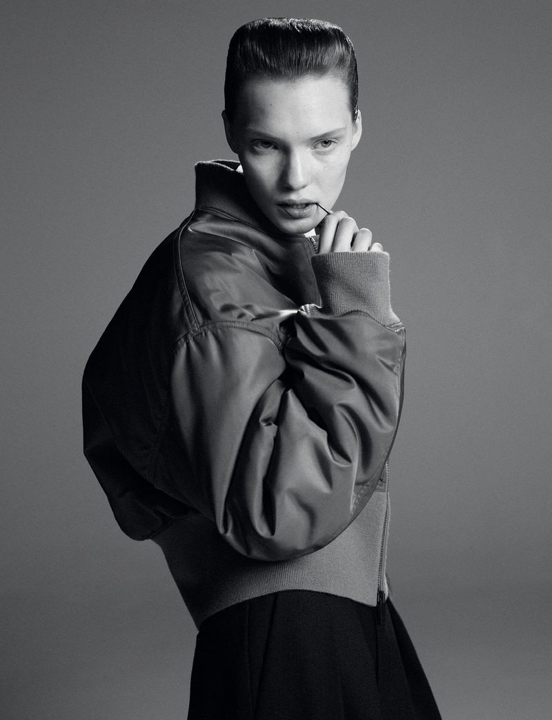 Monochromatic portrait of a young woman with a serious expression, wearing a bomber jacket against a grey backdrop.