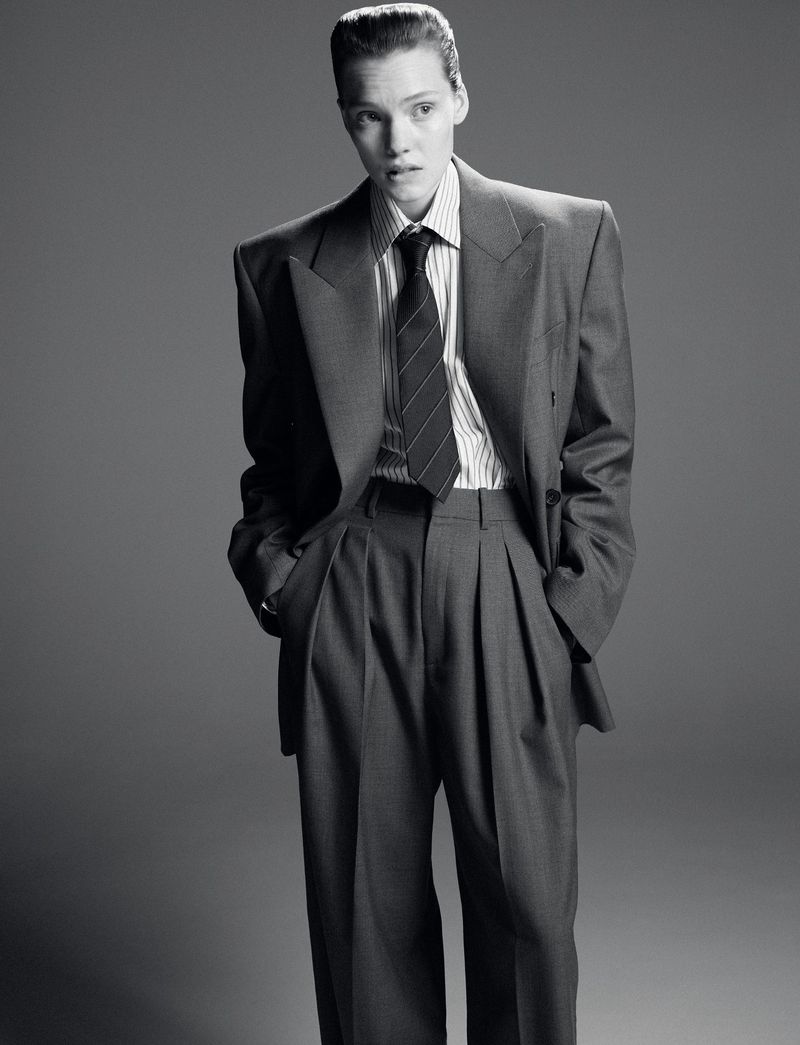 Monochrome portrait of a person in a grey suit, striped shirt, and tie against a grey backdrop, hands in pockets. Fashion photography.
