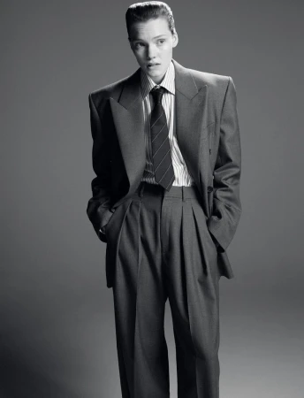 Monochrome portrait of a person in a grey suit, striped shirt, and tie against a grey backdrop, hands in pockets. Fashion photography.