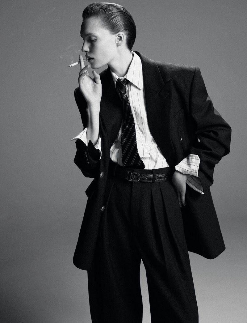 Monochrome portrait of a young adult smoking a cigarette, wearing a suit with a striped shirt and tie, against a neutral gray background.