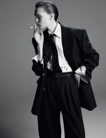Monochrome portrait of a young adult smoking a cigarette, wearing a suit with a striped shirt and tie, against a neutral gray background.